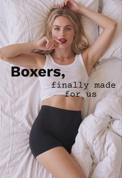 Video of a woman lying on white bed sheets wearing black cotton rich boxer shorts and a white ribbed tank, with the text "Boxers, finally made for us"